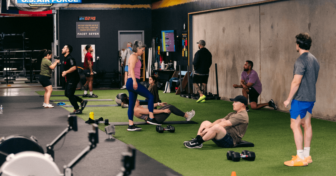 People exercising in a gym with turf flooring, dumbbells, and a workout timer displaying 02:49 on the wall.