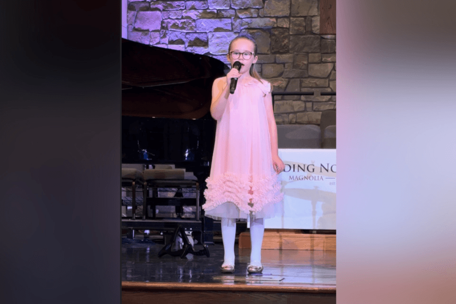 Young girl in pink dress and glasses sings into a microphone on stage near a grand piano.
