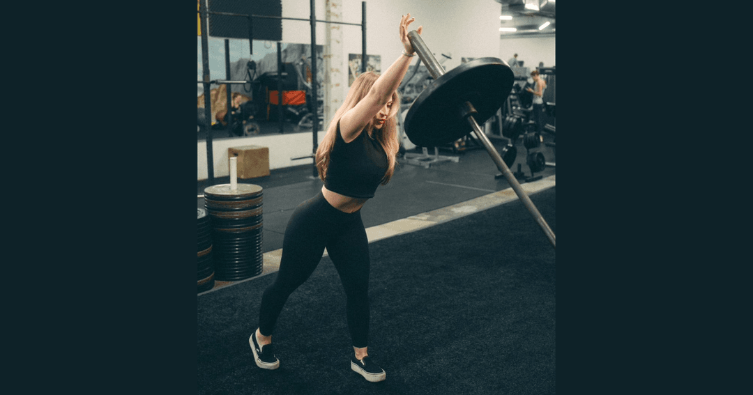 Woman in black workout attire performs a landmine exercise with a weighted barbell in a gym.