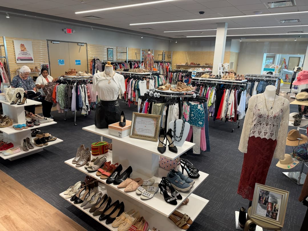 Interior of a thrift or consignment store with clothing racks, shoe displays, mannequins, and shoppers browsing.