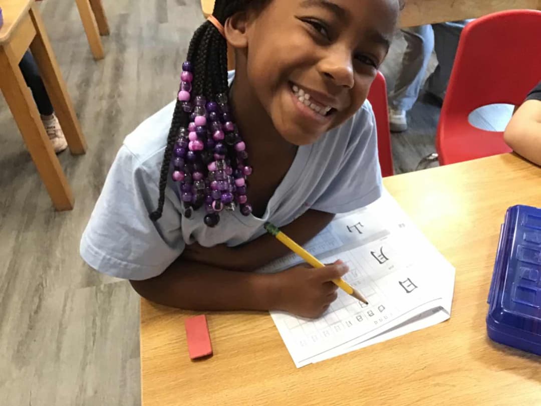 Smiling girl with purple-beaded braids holds a pencil over a worksheet at a classroom desk.