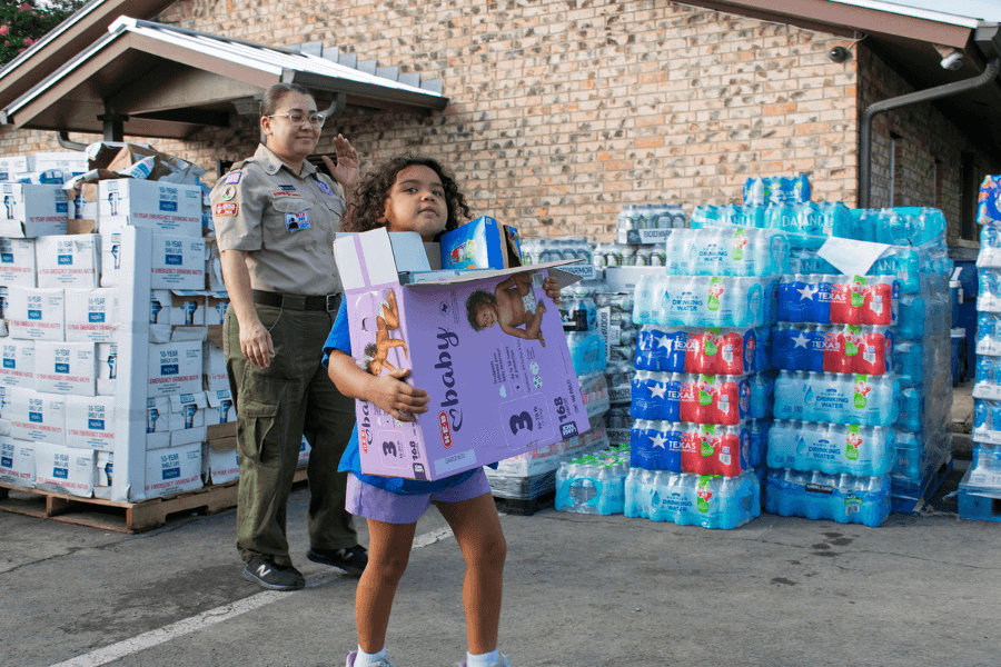 A child carries a box of diapers past pallets of bottled water and emergency supplies at a relief drive.