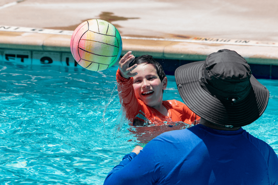 Smiling child in pool reaches for a colorful ball tossed by an adult wearing a sun hat.