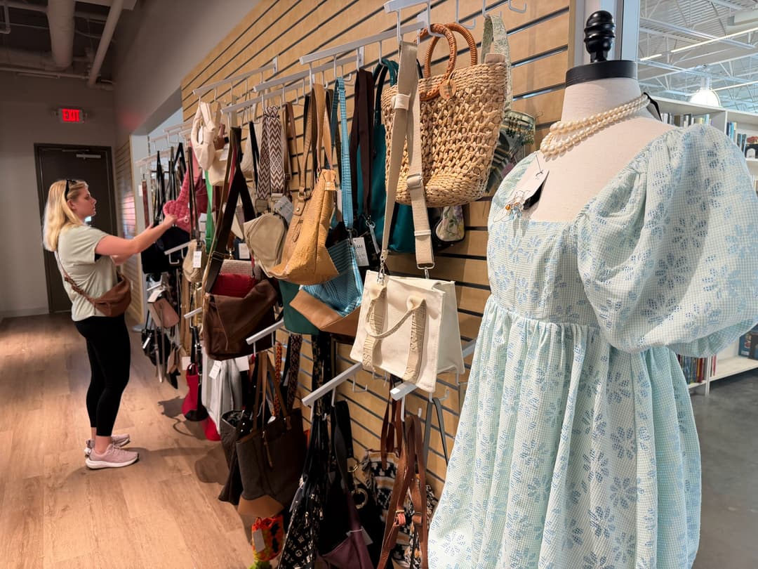 Woman browses handbags on a store wall display beside a mannequin in a blue floral dress with pearls.