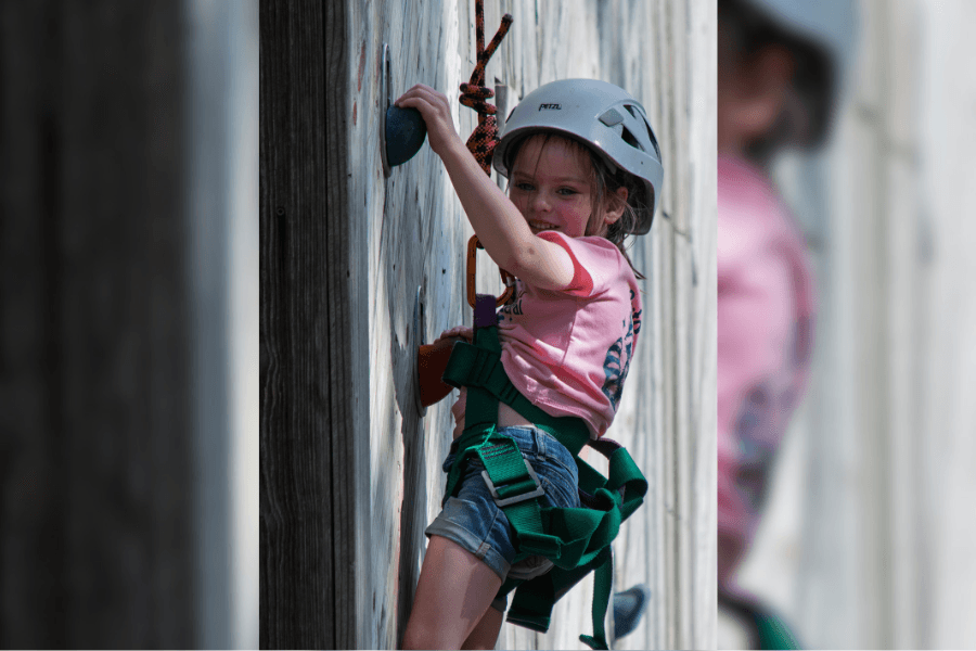 Smiling child in helmet and safety harness reaches for a hold on a climbing wall.