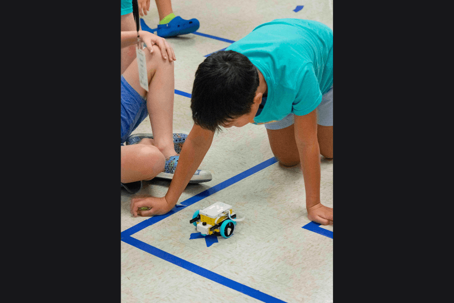 Child in teal shirt watches a small robot follow a blue tape line on the floor.