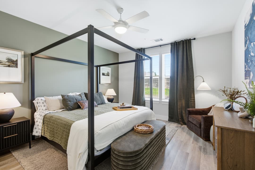 Furnished bedroom with black canopy bed, ceiling fan, sage walls, window with gray curtains, and armchair.