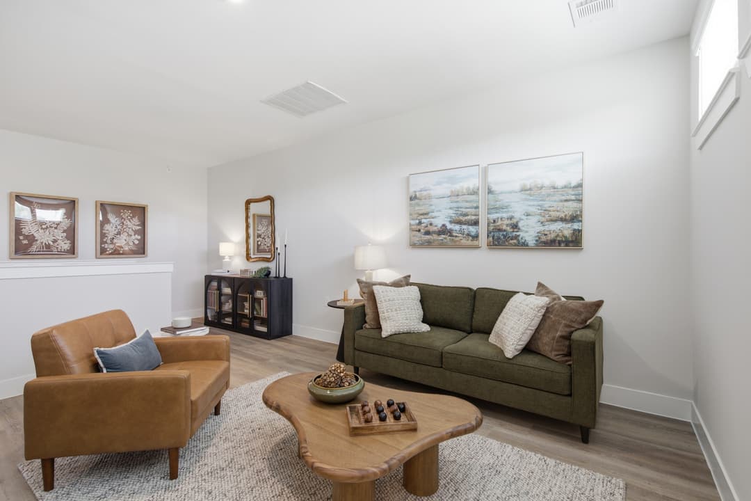 Staged living room with green sofa, tan leather chair, wood coffee table, and framed landscape artwork on white walls.