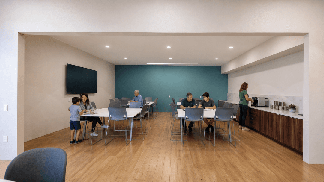 People sit at tables in a modern community room with teal accent wall, wall-mounted TV, and coffee station.