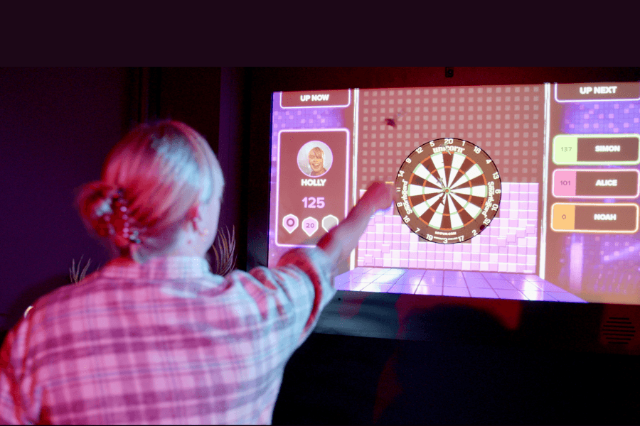 Woman throws a dart at an electronic dartboard display showing scores for players Holly, Simon, Alice and Noah.