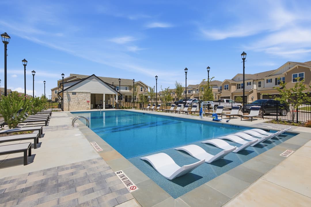 Outdoor pool with in-water lounge chairs, poolside loungers, and townhomes under a blue sky at an apartment community.