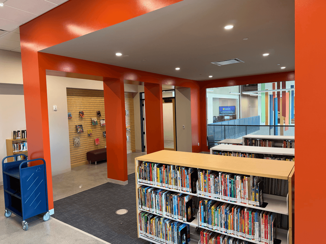 Modern library interior with orange walls, low bookshelves filled with books, a blue cart, and a book display area.