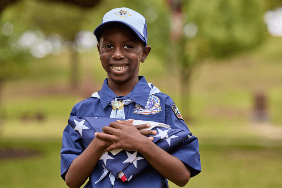 Smiling boy in blue Cub Scout uniform and cap holds a folded American flag outdoors.