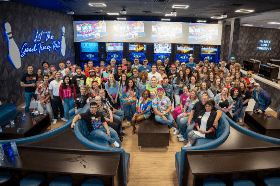 Large group in 80s-themed outfits posing at a bowling alley with "Let The Good Times Roll" neon sign.