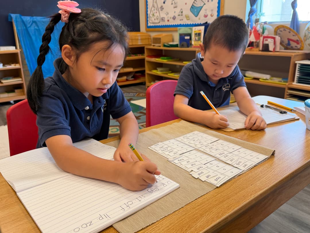 Two young children in navy uniforms write with pencils on lined paper using word cards at a classroom table.