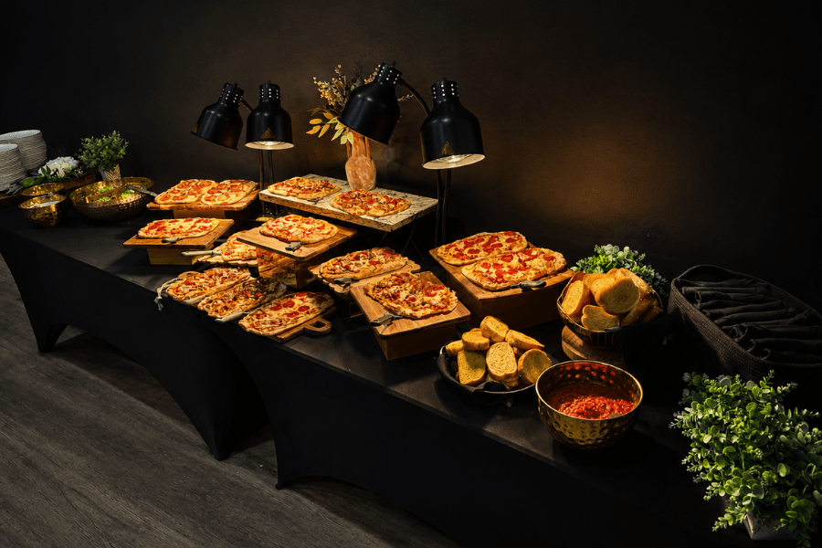 Buffet table with pizzas on wooden boards, bread, dipping sauce, heat lamps and greenery on black cloth.