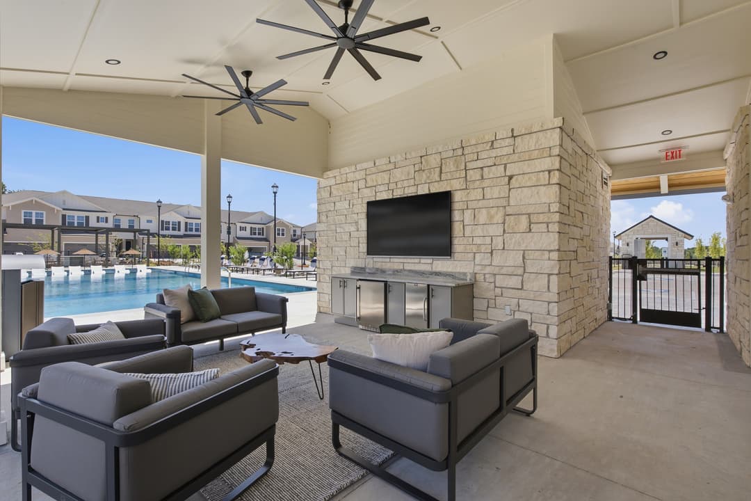 Covered outdoor lounge with gray sofas, wall-mounted TV on stone, ceiling fans, and pool with townhomes beyond.