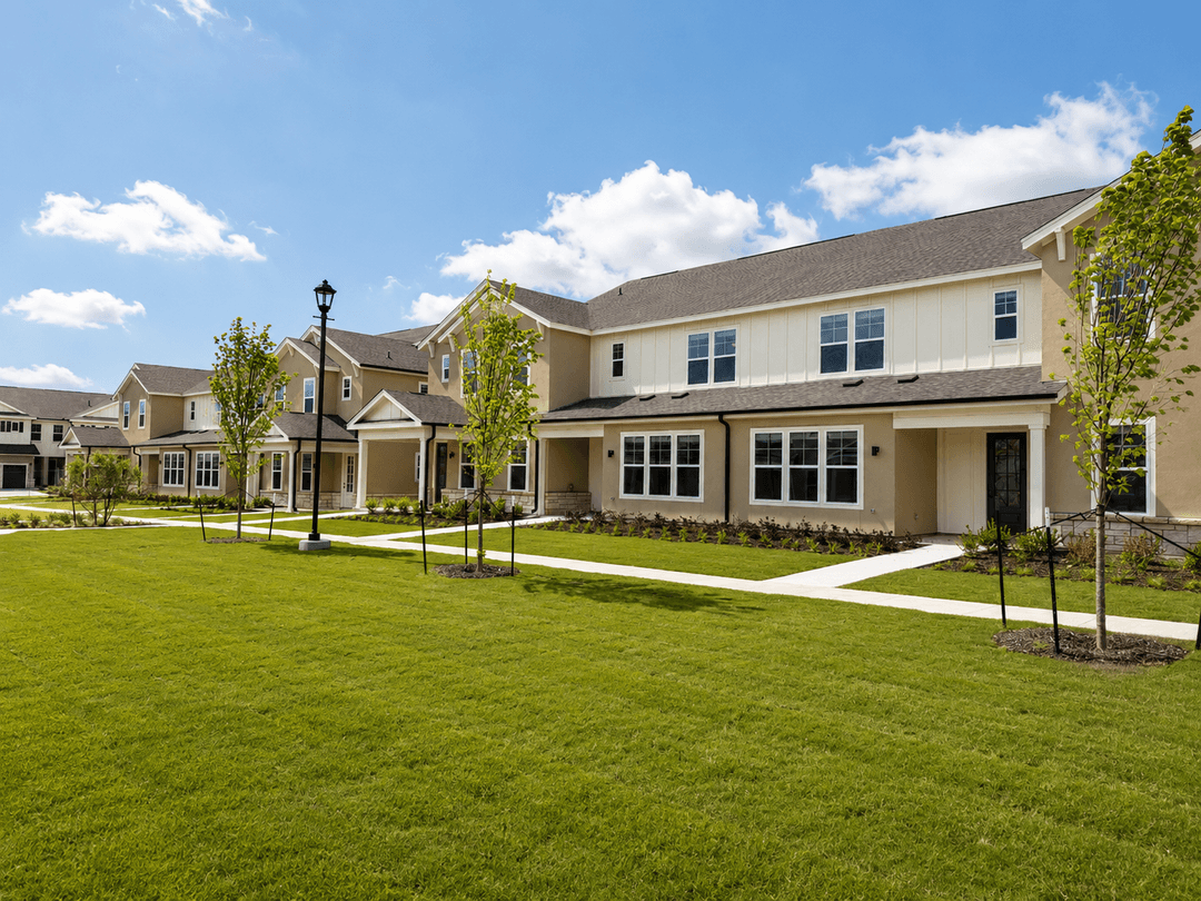 Row of new tan townhomes with manicured green lawn, young trees, and walkways under a blue sky.