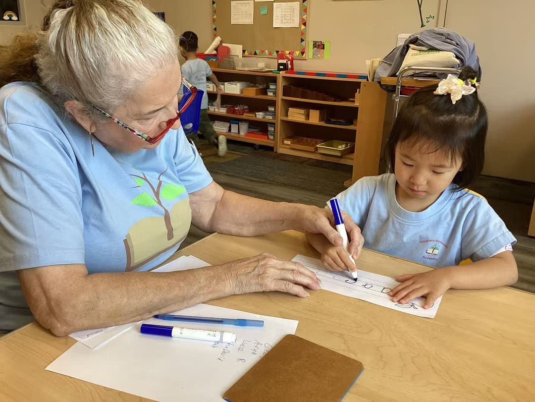 Teacher helps young girl practice writing letters with a marker at a Montessori classroom table.