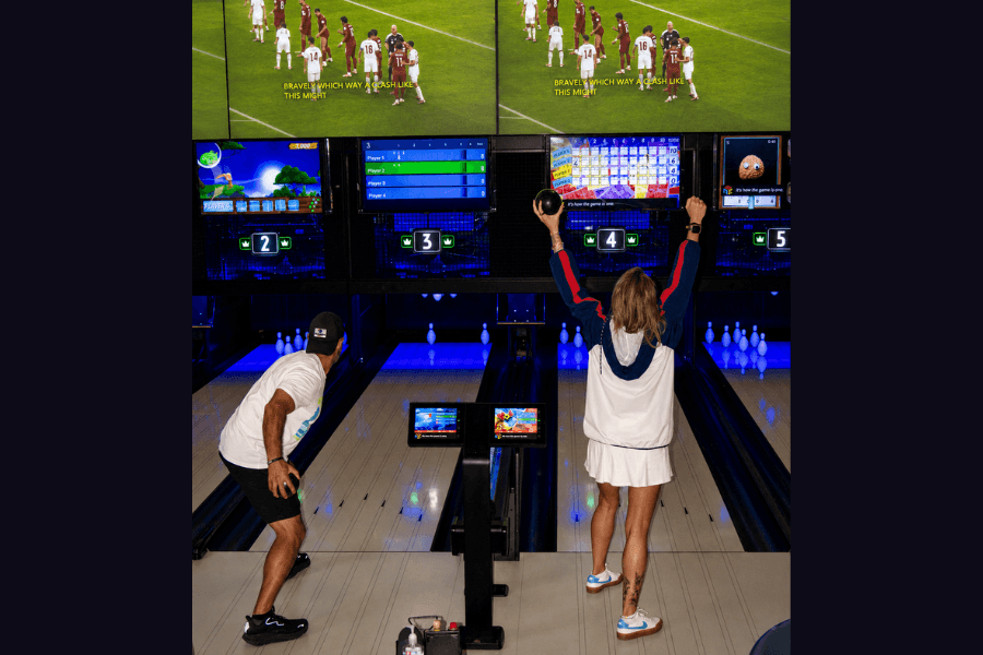 Two people bowling at a neon-lit alley, a woman raising her arms in celebration below screens showing scores and sports.