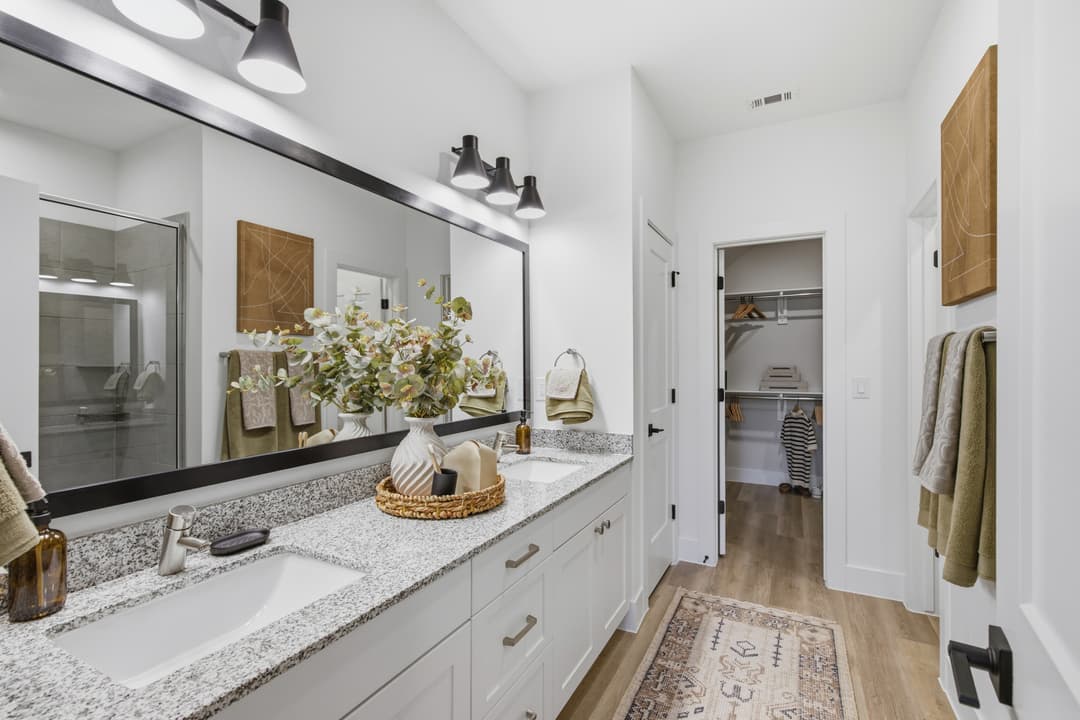 Modern bathroom with double granite vanity, large mirror, glass shower, and adjacent walk-in closet.