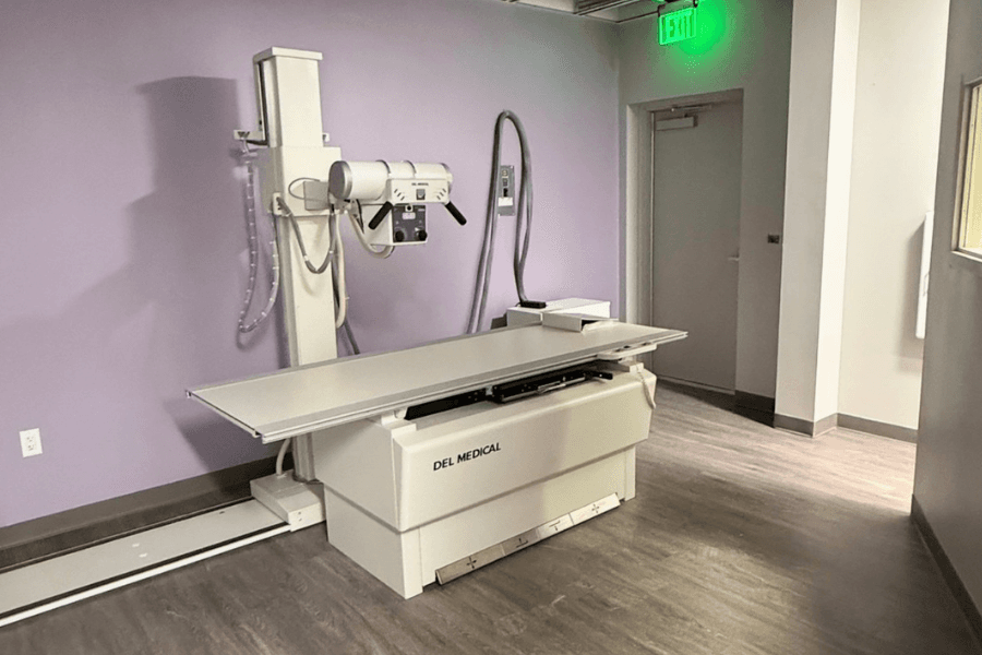 DEL Medical X-ray machine and exam table in a room with purple walls and green exit sign.