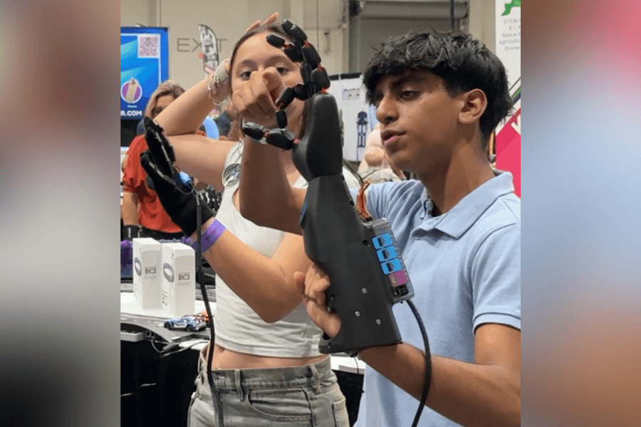 Student holds a black robotic prosthetic hand while a person wearing a sensor glove controls it at a STEM expo.