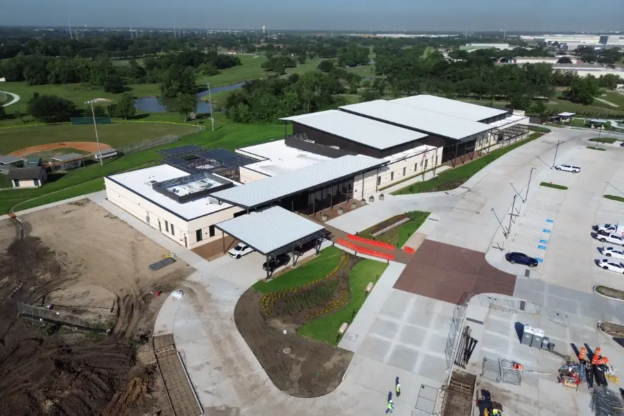 Aerial view of a large modern building with solar panels, parking lots, and construction underway amid green fields.
