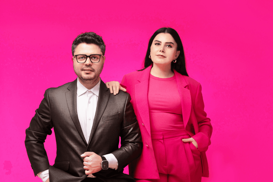Man in dark suit and woman in pink suit pose against a bright pink background.