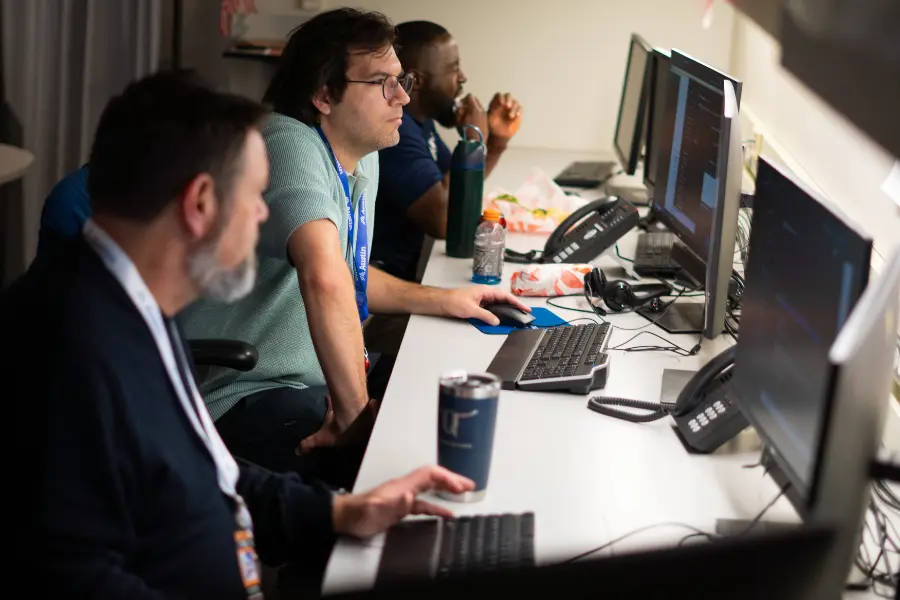 Three men work at computers along a desk with monitors, keyboards, phones, and drinks in a dim office.