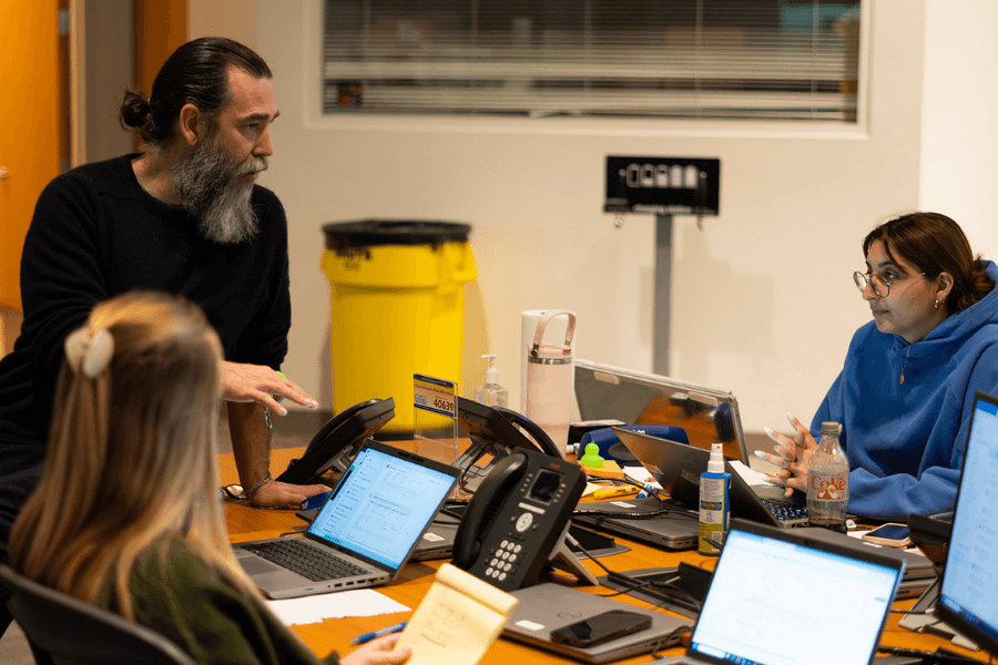 Bearded man leans over table talking to colleagues at a workspace with laptops, phones, and papers.