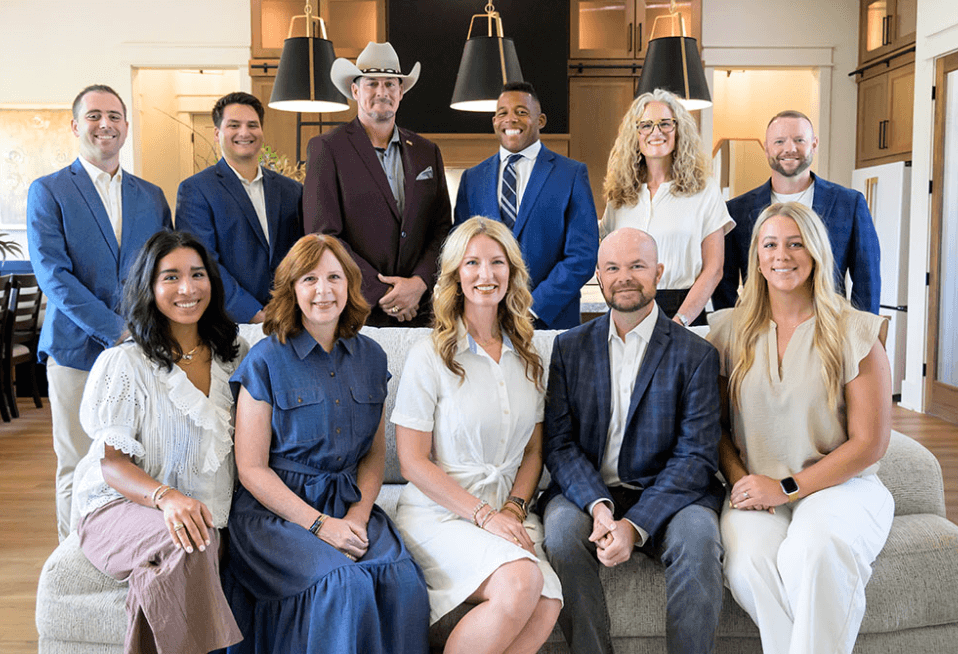 Group of eleven business professionals posing together, five seated on a couch and six standing behind, indoors.