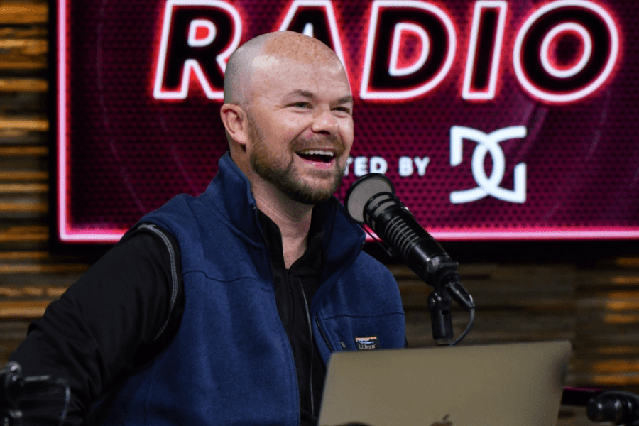 Smiling man in a vest speaks into a microphone at a radio studio with a neon "RADIO" sign behind him.
