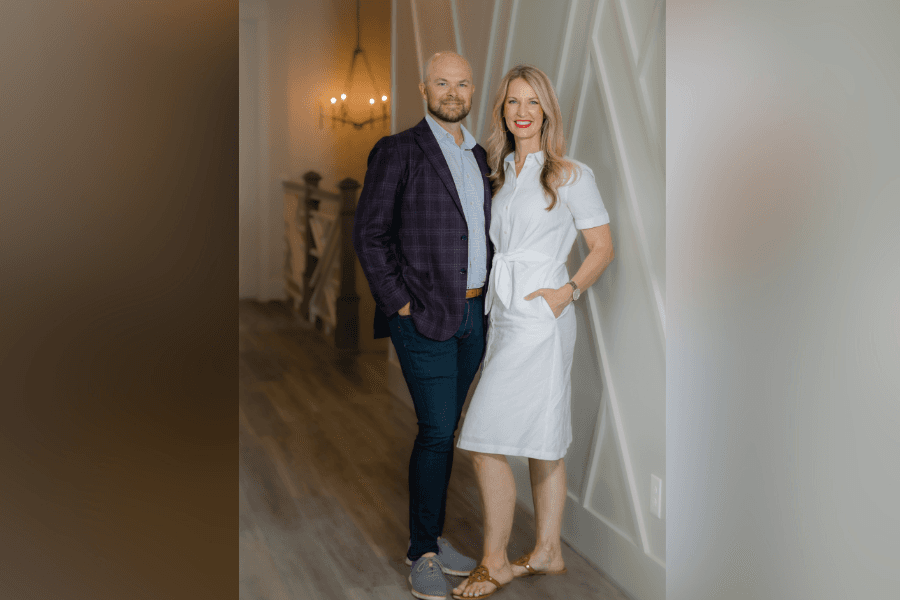 Man in plaid blazer and woman in white dress stand smiling together in a modern home interior.