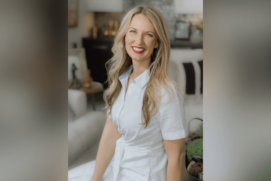 Smiling blonde woman in white shirt dress and red lipstick sits in a well-appointed living room.