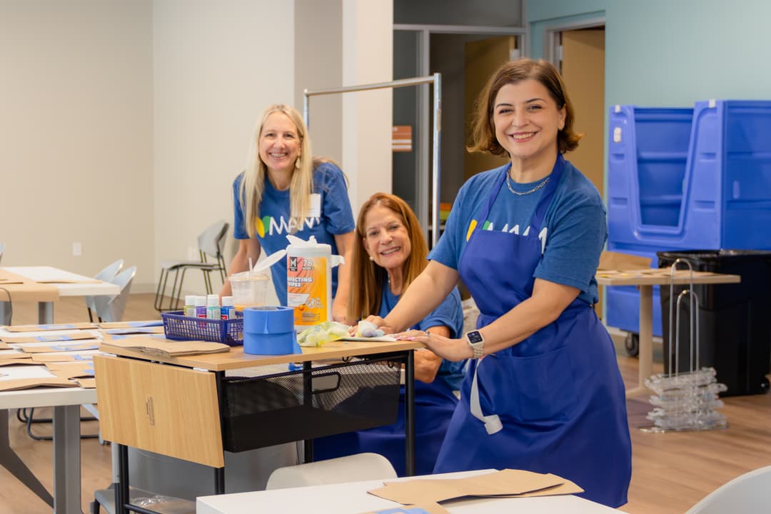 Three smiling women in matching blue volunteer shirts work at a craft table with paint and supplies.