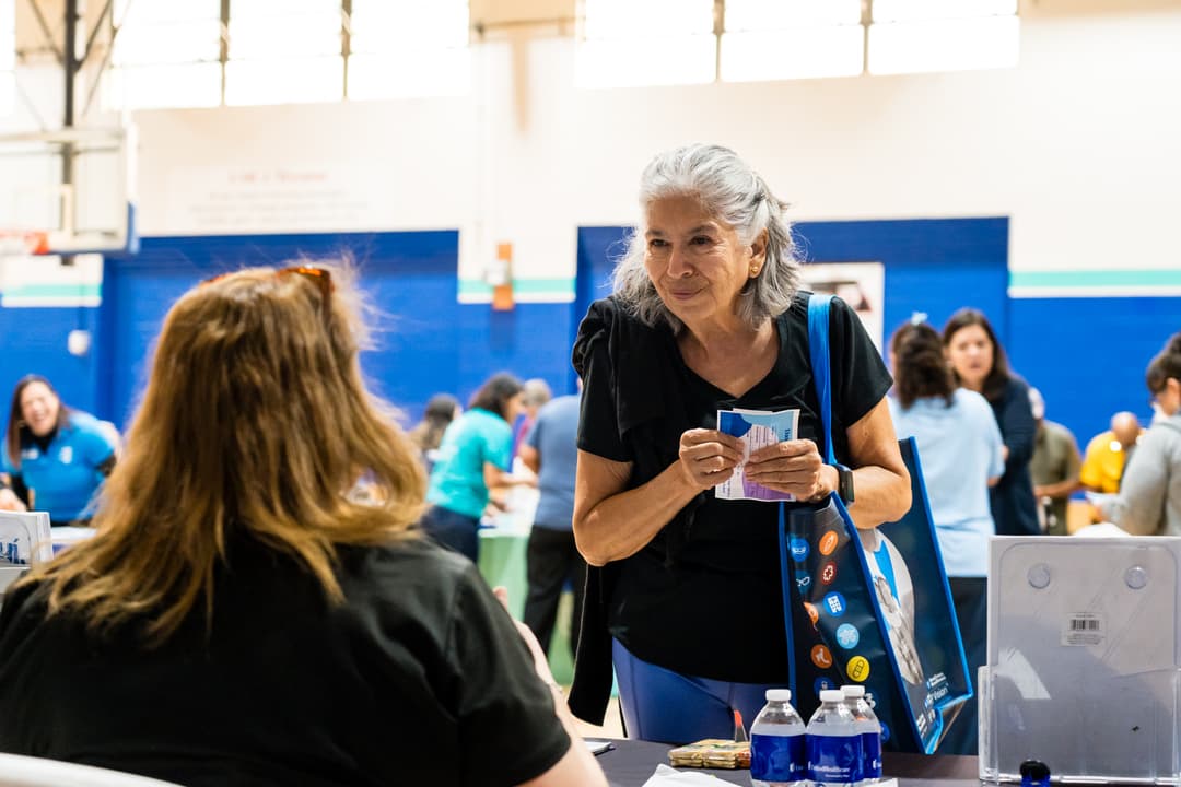 Older woman with gray hair holding a flyer and tote bag talks to a staff member at a health fair information table.