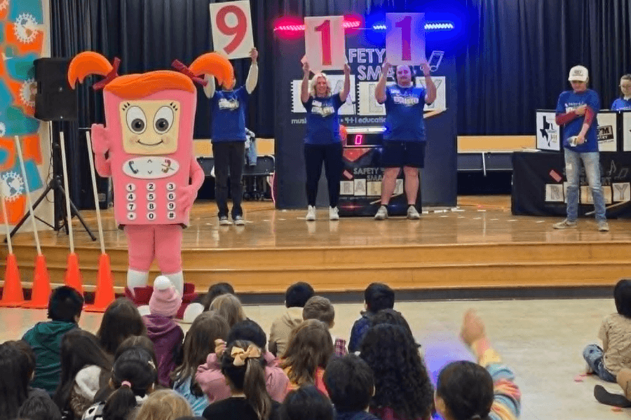 Person in pink phone mascot costume performs on stage before seated children as adults hold up 911 signs.