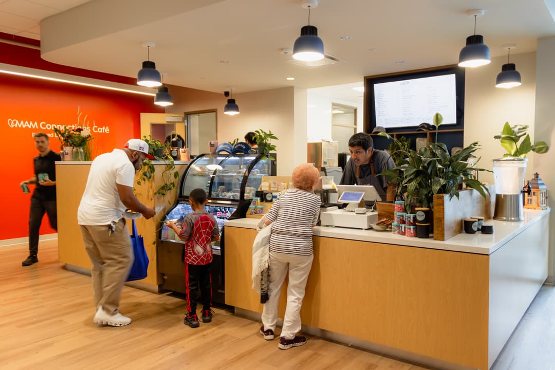 Customers at MAM Connections Café counter as a barista assists an elderly woman; a man and child view the pastry case.