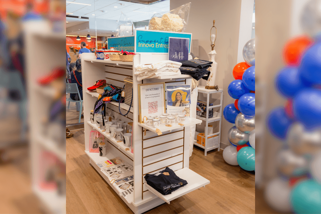 Retail display shelf with T-shirts, mugs and crafts, "Support Our Innova Entrepreneurs" sign, near balloon column.