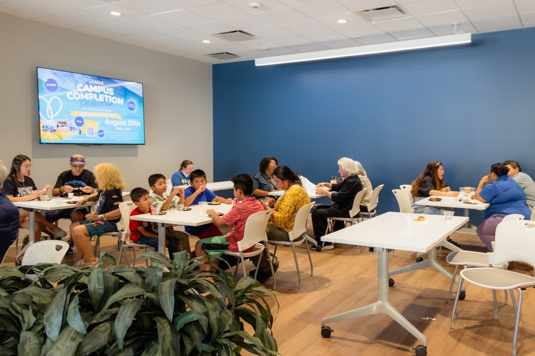 Families sit at tables eating in a room with a screen showing "MAM Campus Completion Celebration, August 29th.