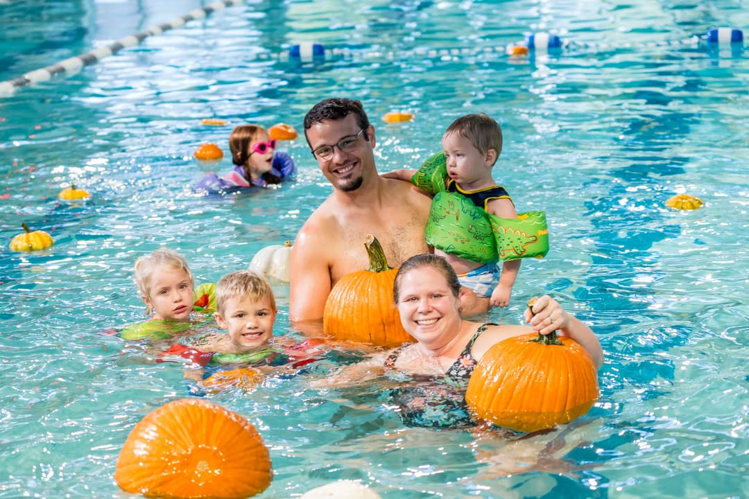 Smiling family with three children in a pool with floating pumpkins at an autumn swim event.