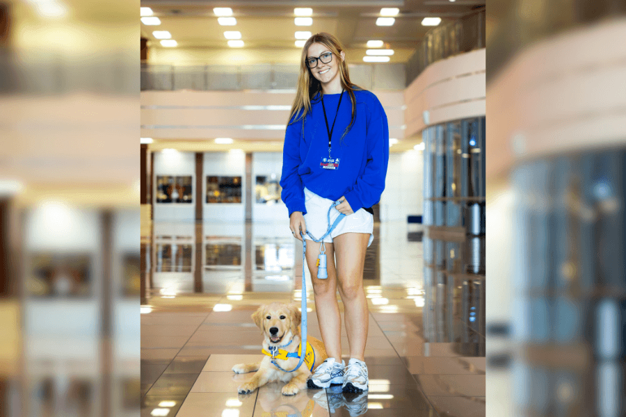 Smiling woman in blue sweatshirt stands with golden retriever service dog puppy in yellow vest indoors.