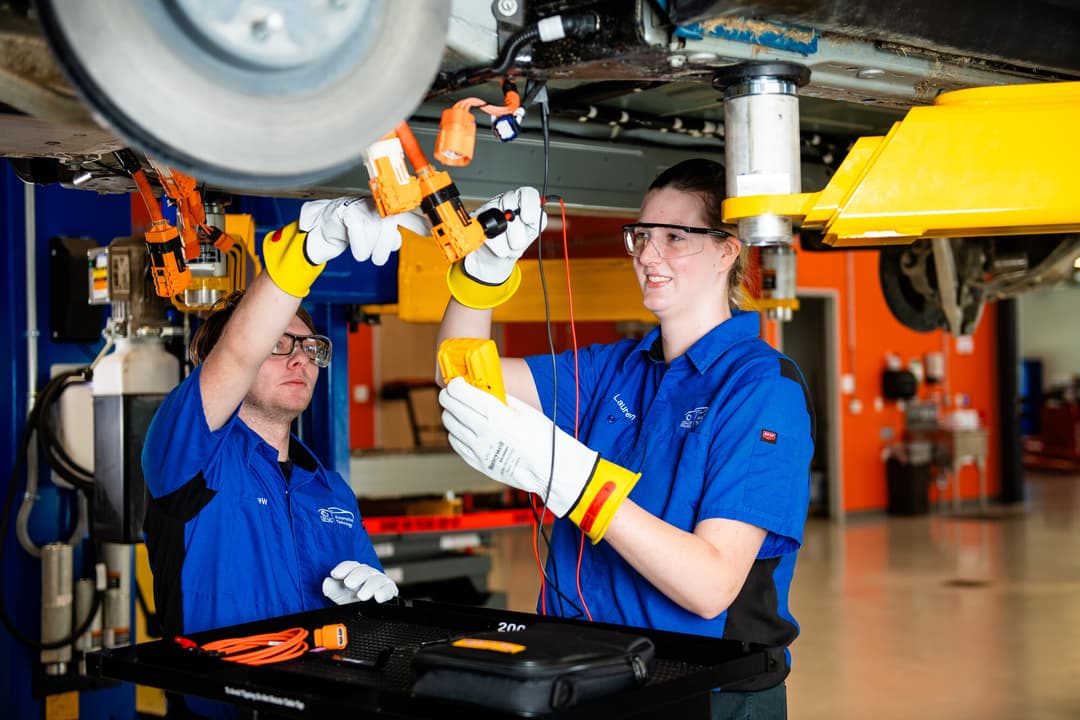 Two automotive technology students in blue shirts and safety gloves work under a lifted vehicle with high-voltage connectors.
