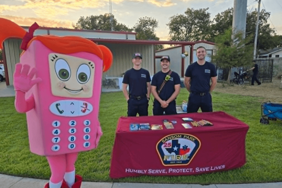 Pink phone mascot beside three firefighters at a Sansom Park Fire outreach table with brochures.