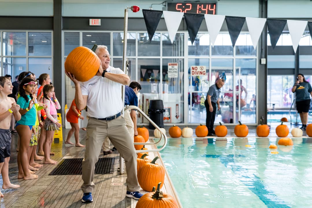 Man in polo shirt hoists a large pumpkin at indoor pool as children watch, with pumpkins lining the pool edge.