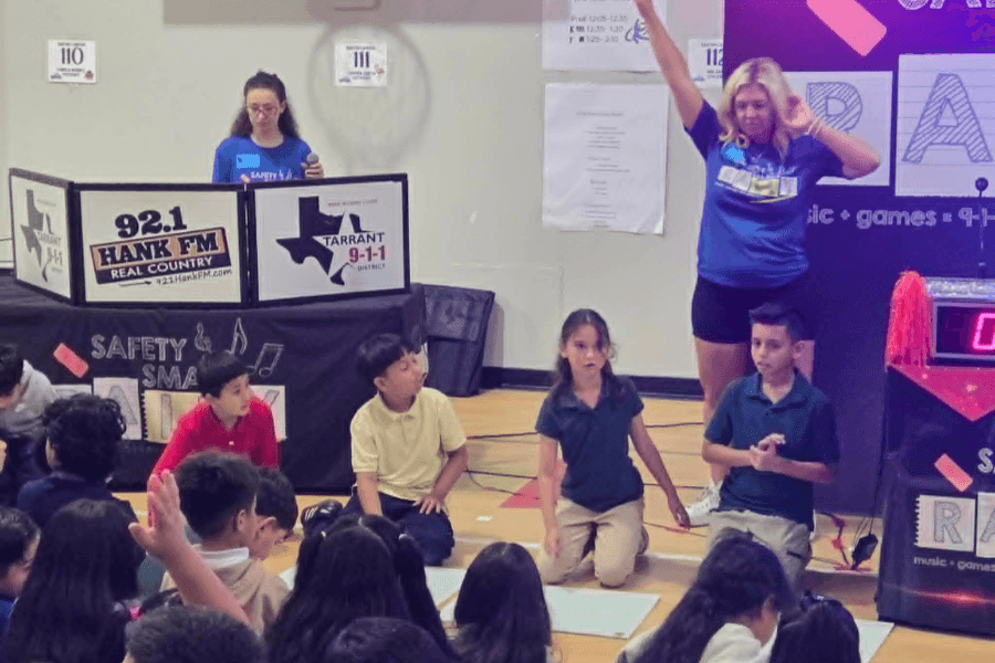 Presenters lead a school safety event for seated children, with 92.1 Hank FM and Tarrant 9-1-1 signage.