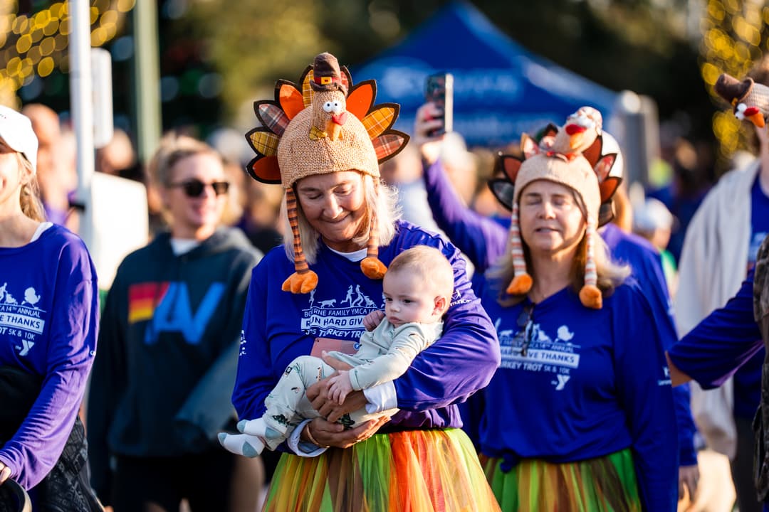 Woman in turkey hat and YMCA Turkey Trot shirt holds a baby, with another turkey-hatted participant beside her.