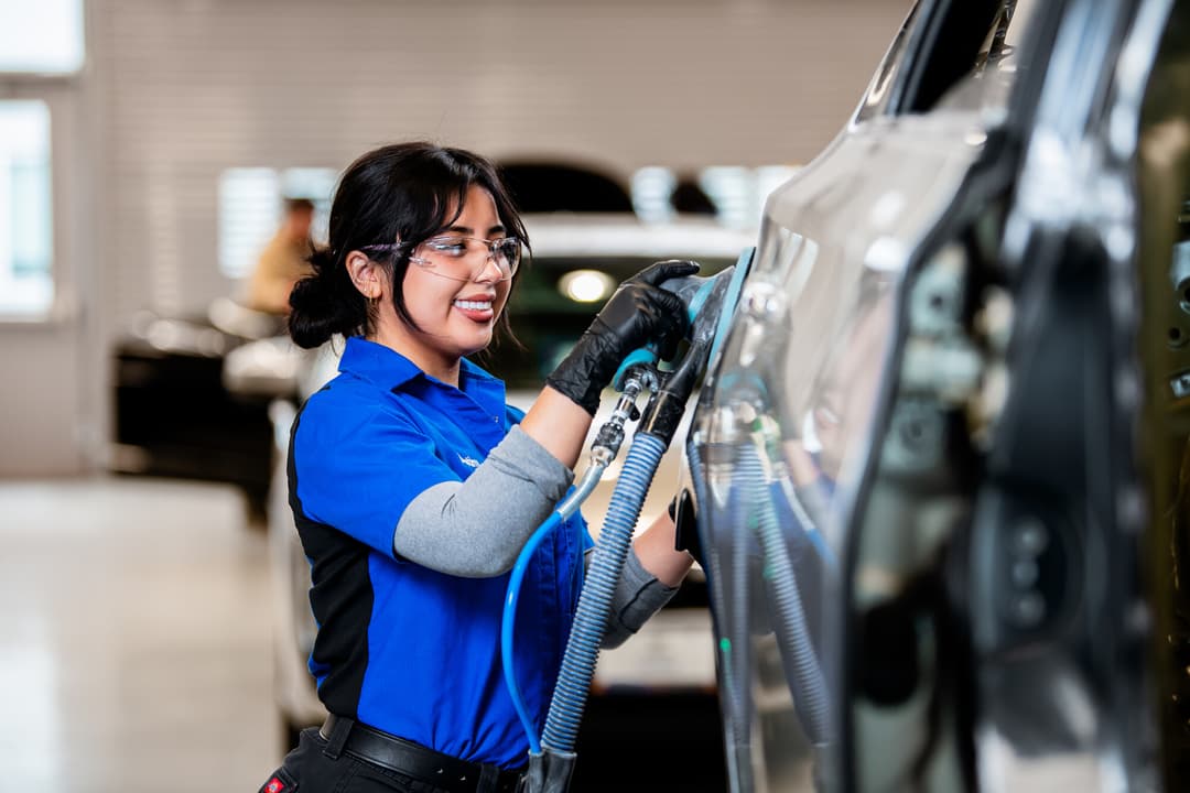 Smiling woman in safety glasses and gloves sands a car panel with a power tool in an auto body shop.