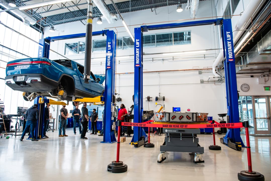 Blue Rivian truck raised on lift as students watch instructor near EV battery behind high-voltage warning barrier in auto shop.